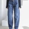 Mujer luciendo jeans baggy azul oscuro con efecto "bigotes" en la parte frontal superior, combinados con blazer cropped negro. Fondo blanco minimalista.