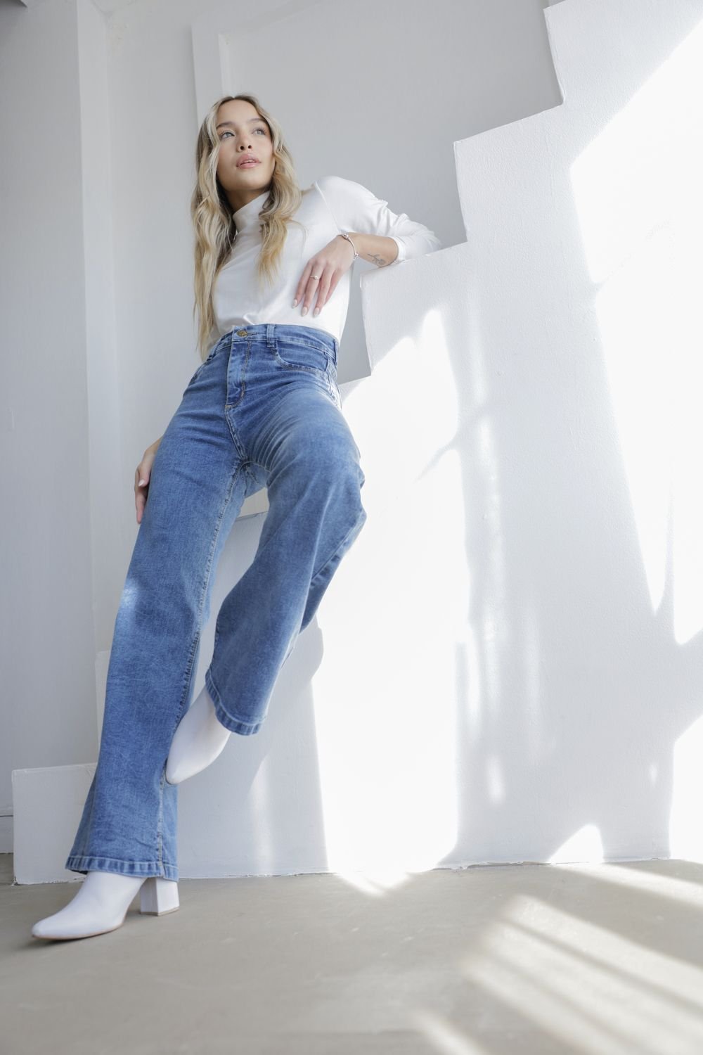 Mujer sentada con jeans wide leg celeste y remera blanca, en escaleras blancas con luz suave.