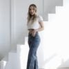Mujer de perfil con jeans wide leg óxido, remera blanca y botas negras, en un ambiente de escaleras blancas.