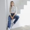 Mujer sentada con jeans mom fit azul clásico, crop top blanco y botas blancas, en ambiente minimalista con luz suave.
