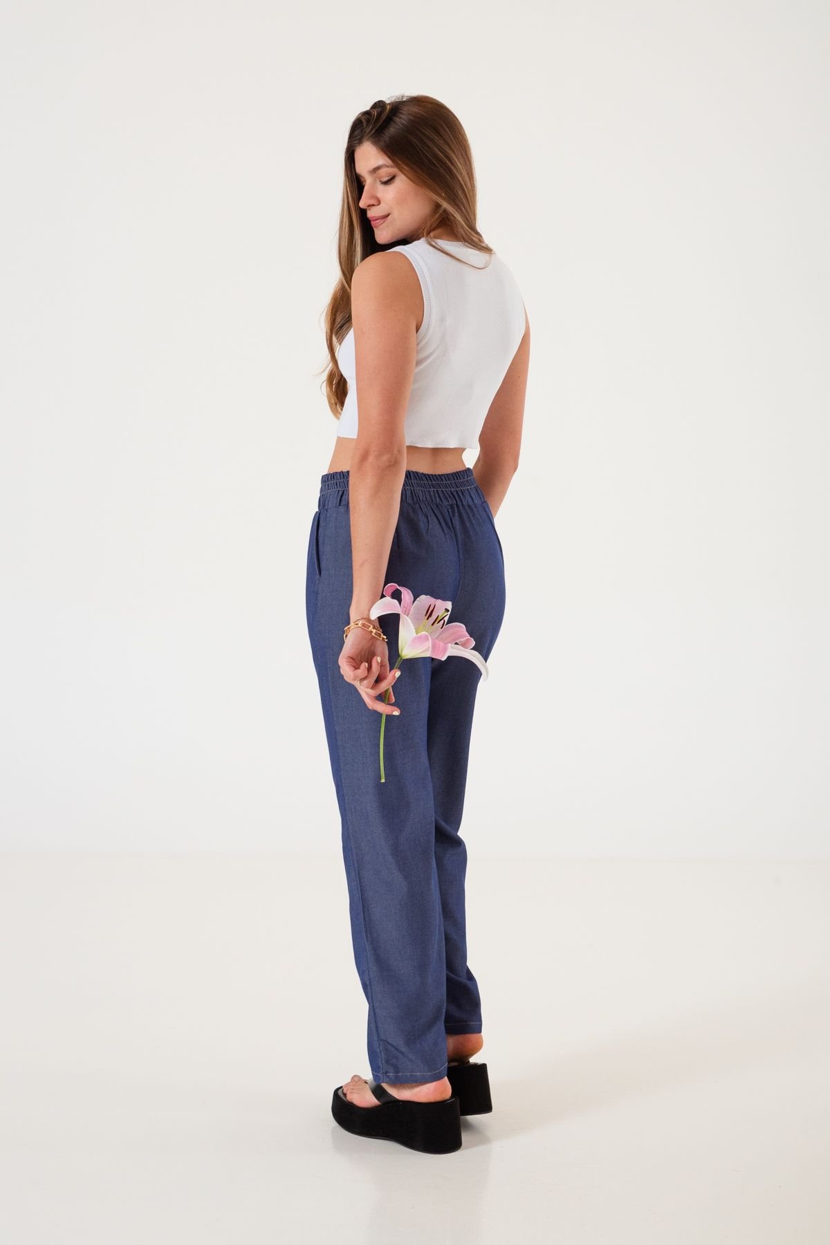 Mujer de espaldas con pantalón recto azul de twill liviano sosteniendo una flor, en ambiente minimalista.